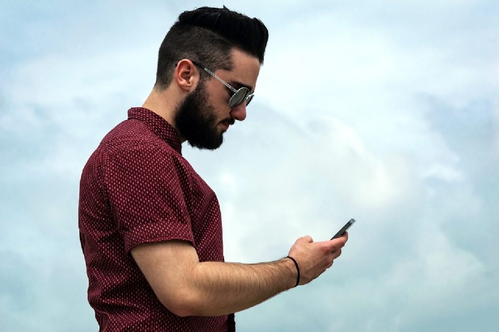 a bearded man holding a cell phone looking down causing what our chiropractor thinks is text neck